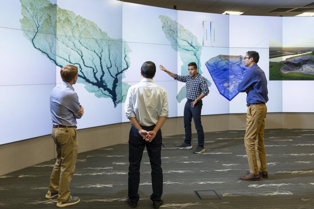This photo is of four men standing in front of a wall of monitors that are showing a tree looking image.