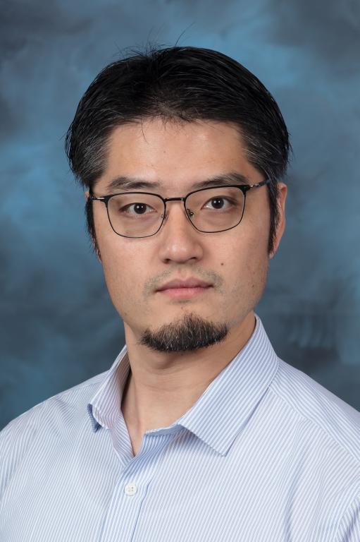 Man with dark brown/black hair with glasses is wearing a light blue button up, smiling for a photo with a blue backdrop.