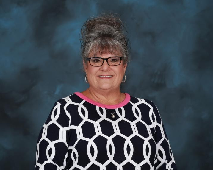 Image is of a woman with grey hair in an updo with black rimmed glasses and a black shirt with white circles and a pink neckline.