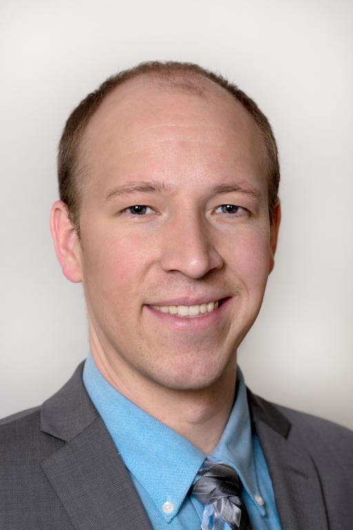 Man in a grey suit and blue button up with a grey tie smiles for a photo with a black backdrop.