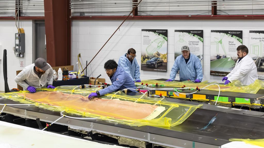 5 scientists in blue and white coats are leaning over the wind blades covered in orange and yellow material