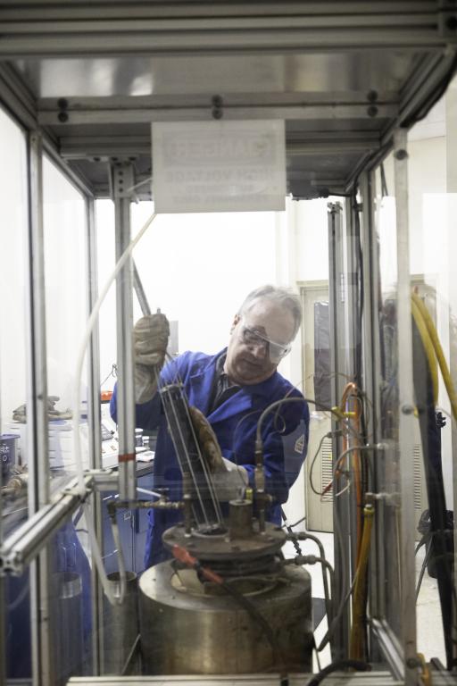 Eric Wolfe loads pellets of coal char into a reactor, wearing blue coat, gloves and safety goggles. 