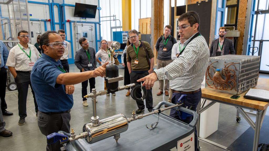 Two scientists are standing in the lab pointing at an object on the table, both wearing glasses.