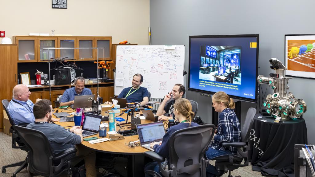 7 researchers are sitting around an oval table talking as a group. There is a whiteboard with writing and a blue screen in the background. 