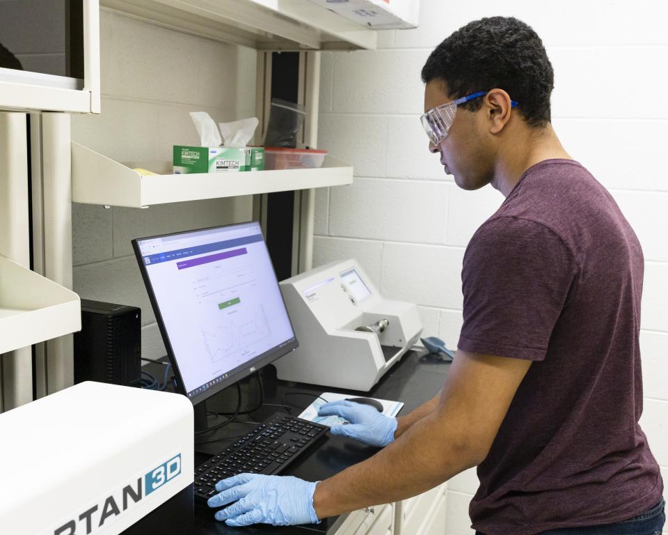 Researcher is standing at his lab's computer with blue gloves and glasses, working on something on the computer