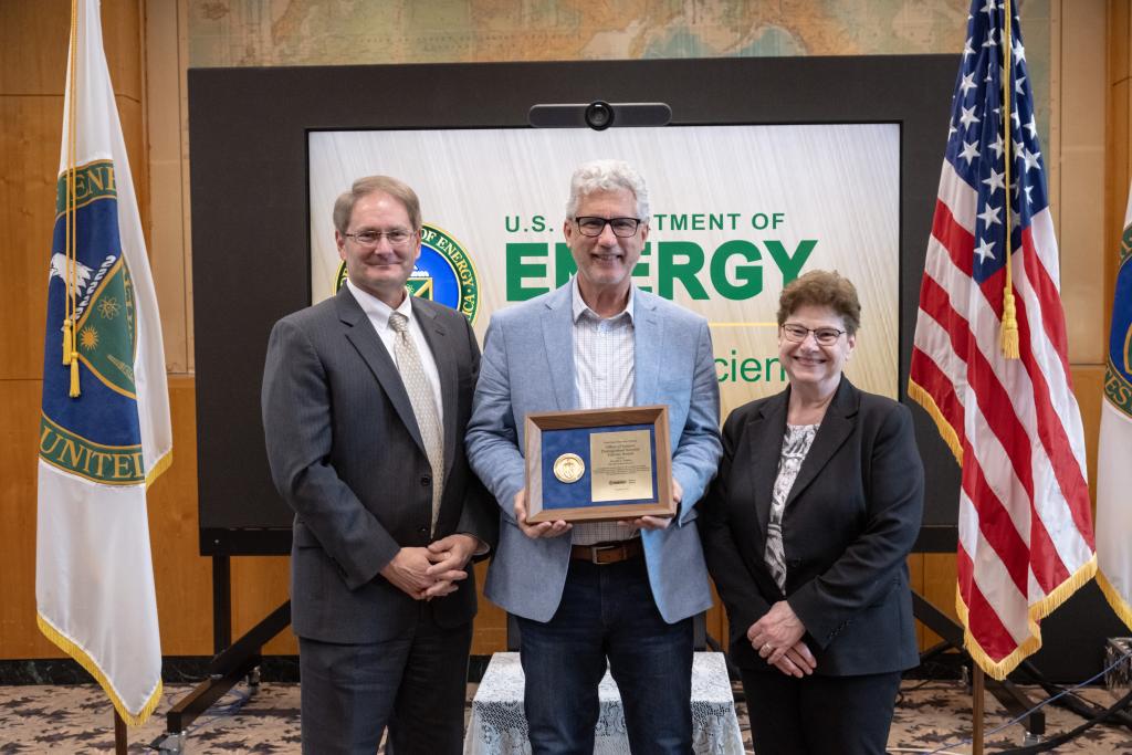 three well-dressed, smiling people stand between a flag with the U.S. Department of Energy seal and the American flag. The person in the center holds an award plaque