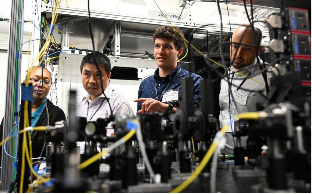 Four scientists are gathered looking at a screen, one in the middle is pointing his figure to motion at what to look at. 