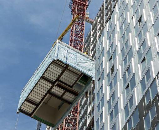 A tall grey building is off to the right, towering over a living space module that is being hoisted by a crane 