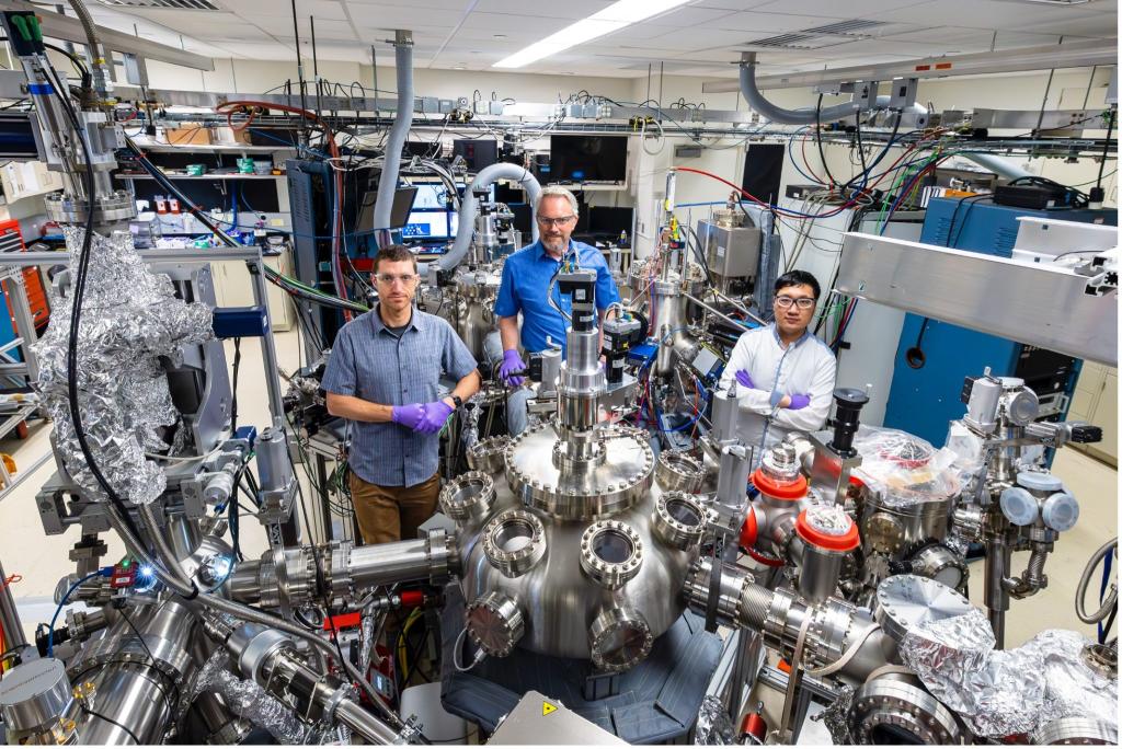 Three researchers are standing in the quantum computing lab at ORNL behind a big metal machine with multiple port hole looking windows attached.