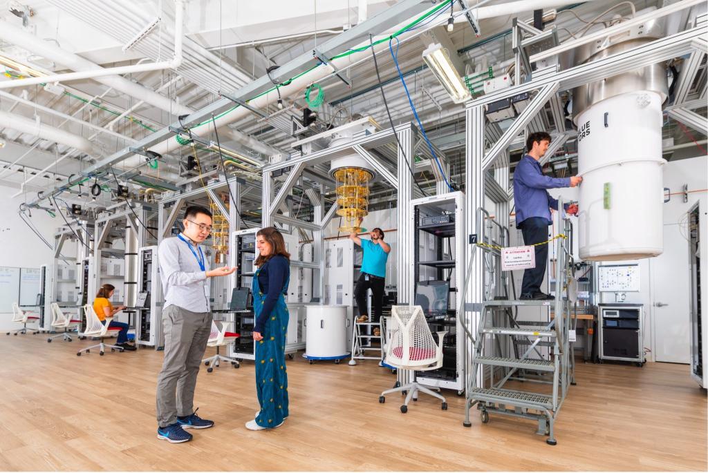 Two researchers are at the SQMS Quantum Garage at Fermilab talking while there are three researchers working on different areas in the background, two standing on ladders