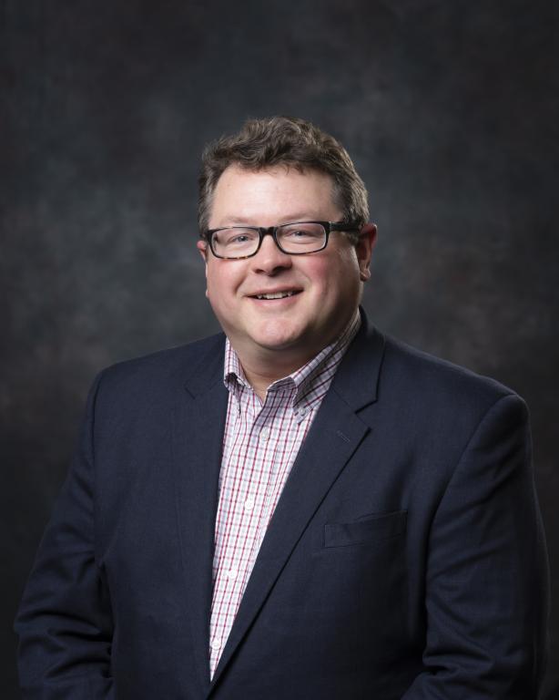 ORNL program manager Bill Peter is posing for a photo with a dark grey background and black suit with glasses