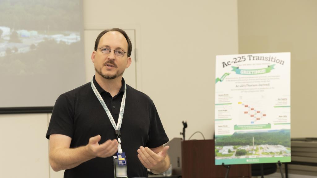 A male researcher is standing next to a poster board displayed on an easel to the right. The researcher is dressed in professional attire, and the poster board is positioned beside him, showing research or visual information.
