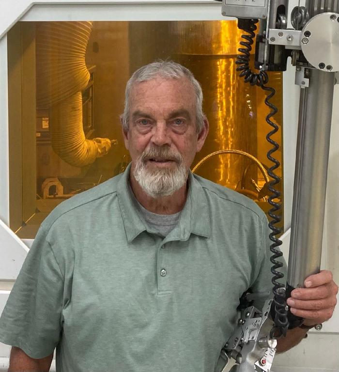 David Dunning, a master technician at ORNL is standing, posing for a photo grabbing a piece of metal equipment. The background is yellow. 