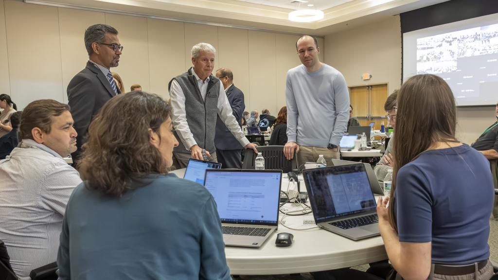 Secretary Wright, center, talks with ORNL participants during the “1,000 Scientists AI Jam Session,” along with ORNL’s Prasanna Balaprakash (left) and OpenAI co-founder Greg Brockman - with the participants backs to camera and their computers on a round table. 
