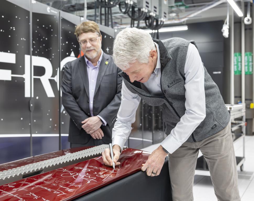 Secretary Wright is bent down signing a red computer cabinet door with silver sharpie while Streiffer looks on. 