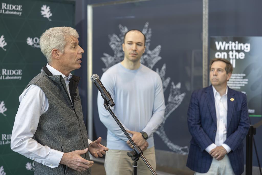 Secretary Wright, left, addresses media, standing up at a microphone with a green backdrop with ORNL's logo "Oak Ridge National Laboratory" and an oak leaf multiplied throughout. There are two men standing off to the right of the speaker. 