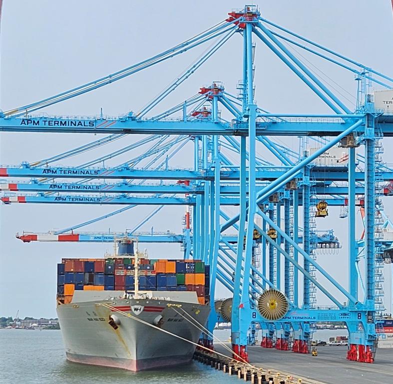 A cargo ship to the left of the seaport with bright blue metal surrounding it