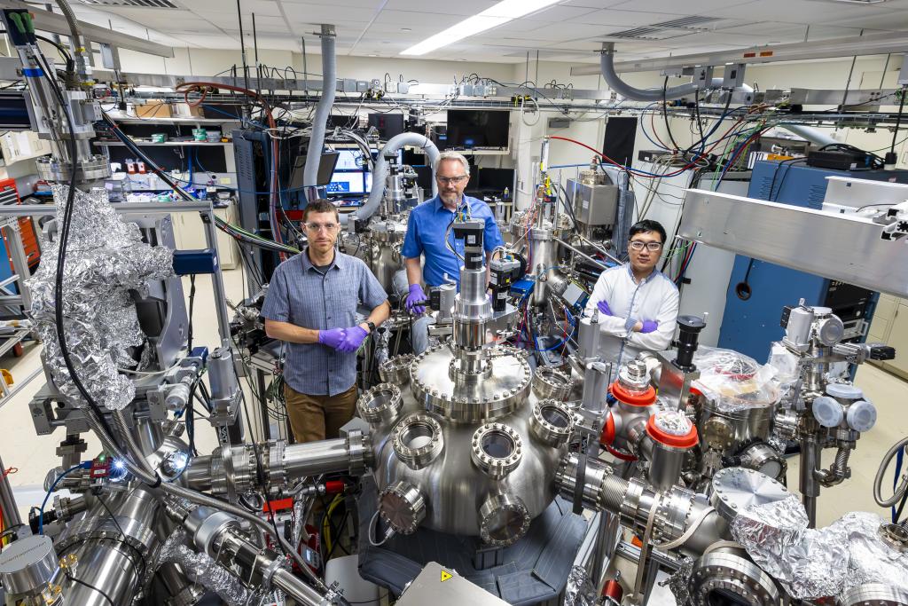 Three scientists are standing in the lab behind the big metal machine with purple gloves on