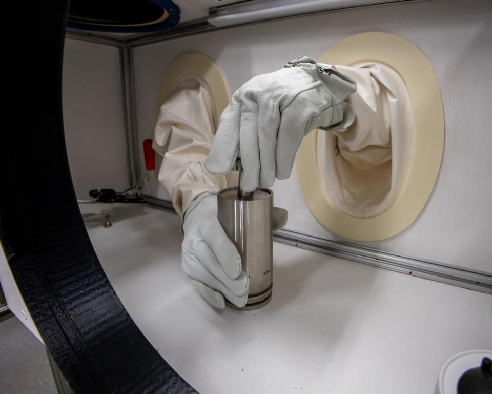Two hands in gloves are holding a silver tube inside a white box with openings for the hands to poke through