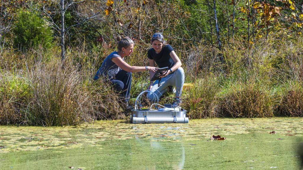 two researchers kneeling at the edge of a body of water covered in algae referencing a table that controls an aquatic drone in the water in front of them