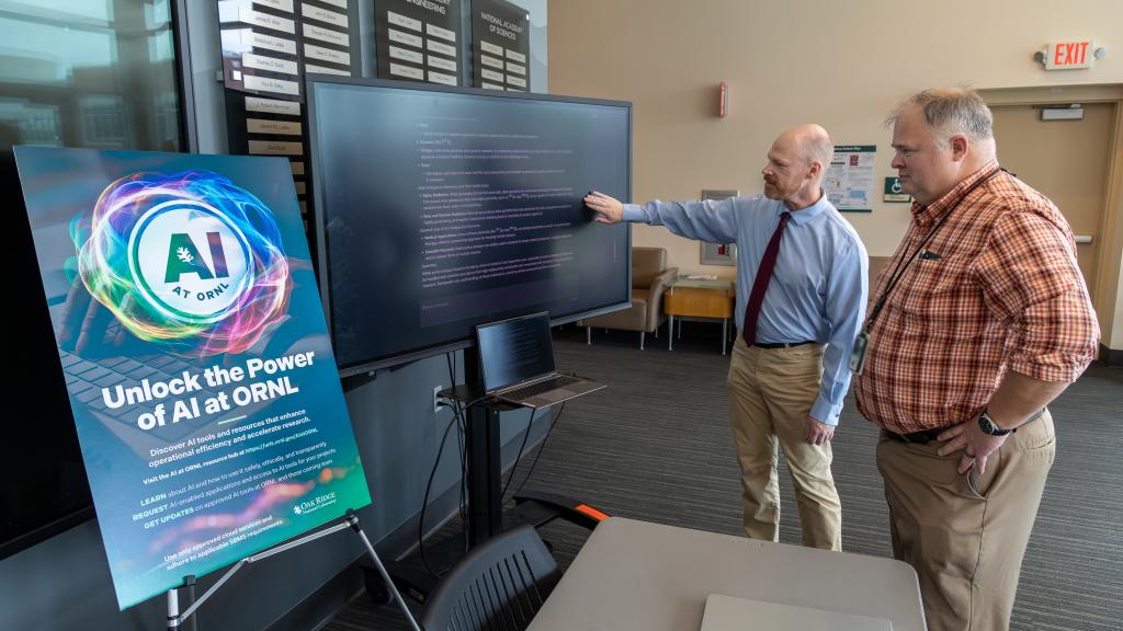 Two ORNL researchers are standing to the right of a computer screen and a poster promoting the AI Expo