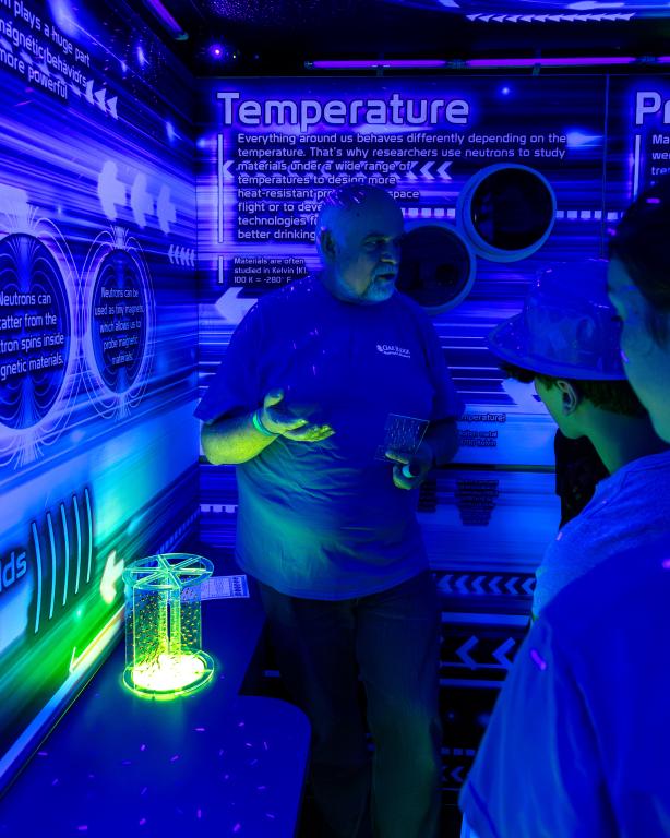 Thomas Proffen walks fourth and fifth graders through ORNL’s Traveling Science Fair. The photo is dark blue with a bright green glow-n-the-dark material to his left