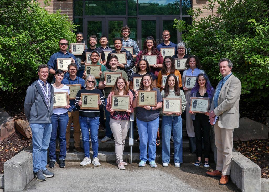group photo taken in front of a brick building of people holding award plaques