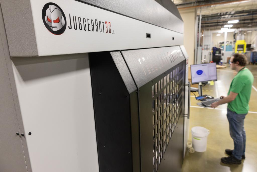 ORNL technical staff member is operating the Juggerbox 3D printer at the MDF. The Juggerbot robot logo is in the foreground of the photo