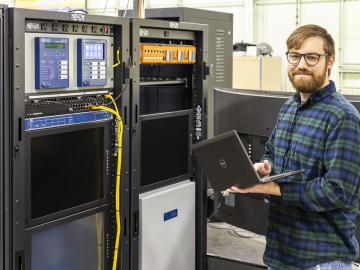 A man wearing glasses and holding a laptop stands near cyber-physical equipment.