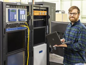 A man wearing glasses and holding a laptop stands near cyber-physical equipment.