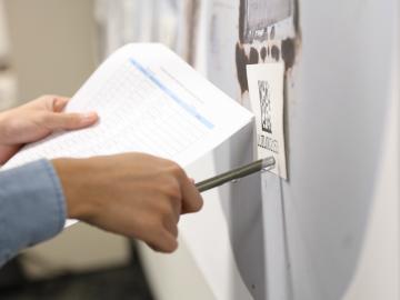 A person holding a pen in their right hand and a paper in their left looks at a sign on the wall. 