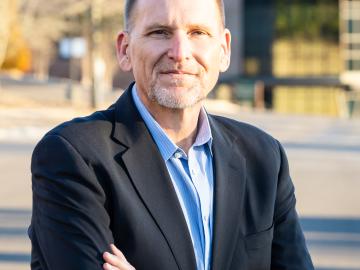 Photo of a man in a black jacket suit with a blue shirt crossing his arms outside. He is smiling.