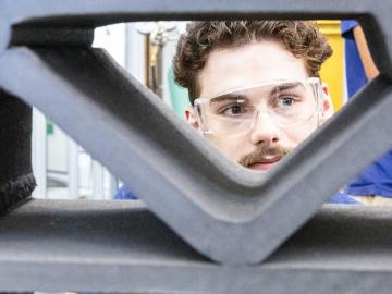 Male student wearing clear safety goggles peering through a 3D-printed item.