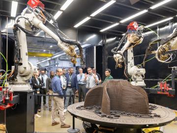 Two robotic arms, one on the left side of the frame and one on the right  - are pointing at a brown three pronged shape on a table. There are people in the background looking at this setup. 