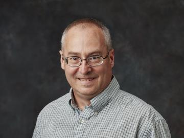 Man in glasses and a collared shirt smiles in front of a black backdrop