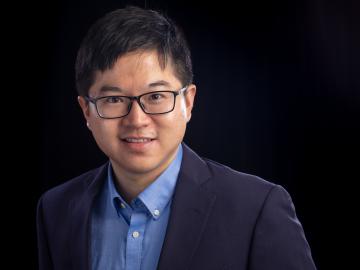 Man in blue suit jacket, blue button down shirt, glasses and dark hair smiles with a black backdrop. 