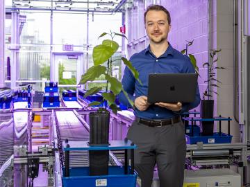 Man in blue shirt and grey pants holds laptop and poses next to a green plant in a lab. 