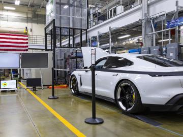 White car (Porsche Taycan) with the hood popped is inside the building with an american flag on the wall.