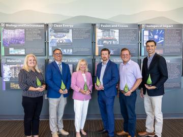 Six people are standing, holding awards, posing in front of a wall with mountain wallpaper. Left to right it goes girl, boy, girl, boy, boy, boy. 