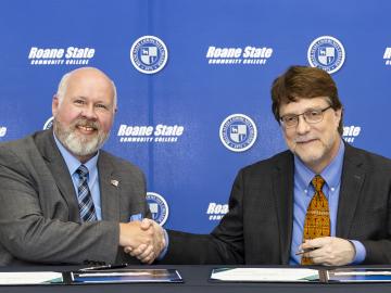 Two men shake hands in front of a blue background. Both are wearing suites and a tie. 