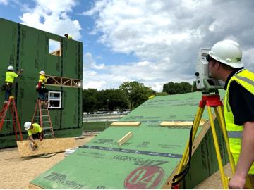 Man in bright yellow safety vest and hard hat is looking through a small machine that is pointed at a house being constructed. 