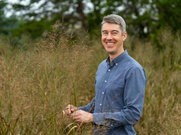 Man in blue button down shirt with short grey hair stands in a field smiling