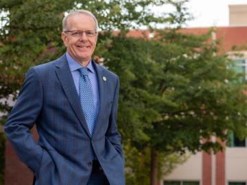 Man in blue suit and tie with glasses and grey hair poses outside in front of trees and a brick building