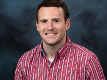 Man in red and white stripe button down shirt with dark brown hair poses for photo in front of blue/grey background.