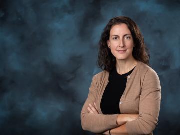 Image is of woman with dark hair crossing her arms and posting for a photo in front of a blue backdrop. She is wearing a black shirt with a tan cardigan. 