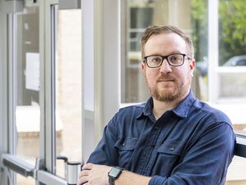 Man is leaning against the window, arms crossed in a dark navy button up. 