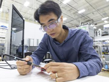 This photo is of a male scientist sitting at a desk working with materials, wearing protective glasses.