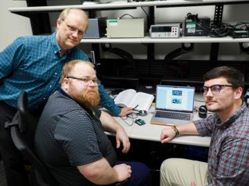 This photo is of three men sitting around a laptop computer that happens to be working on cybersecurity testing equipment. 