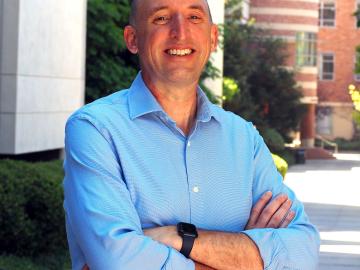Man in blue button down shirt poses outside for a picture with his arms crossed. 