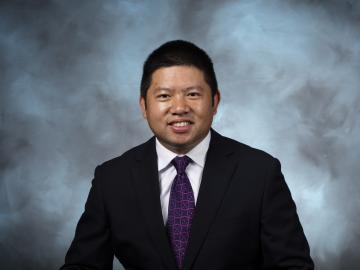 Man with short, dark black hair is wearing a black suit jacket, purple tie and white button down, smiling for a photo in front of a blue grey background